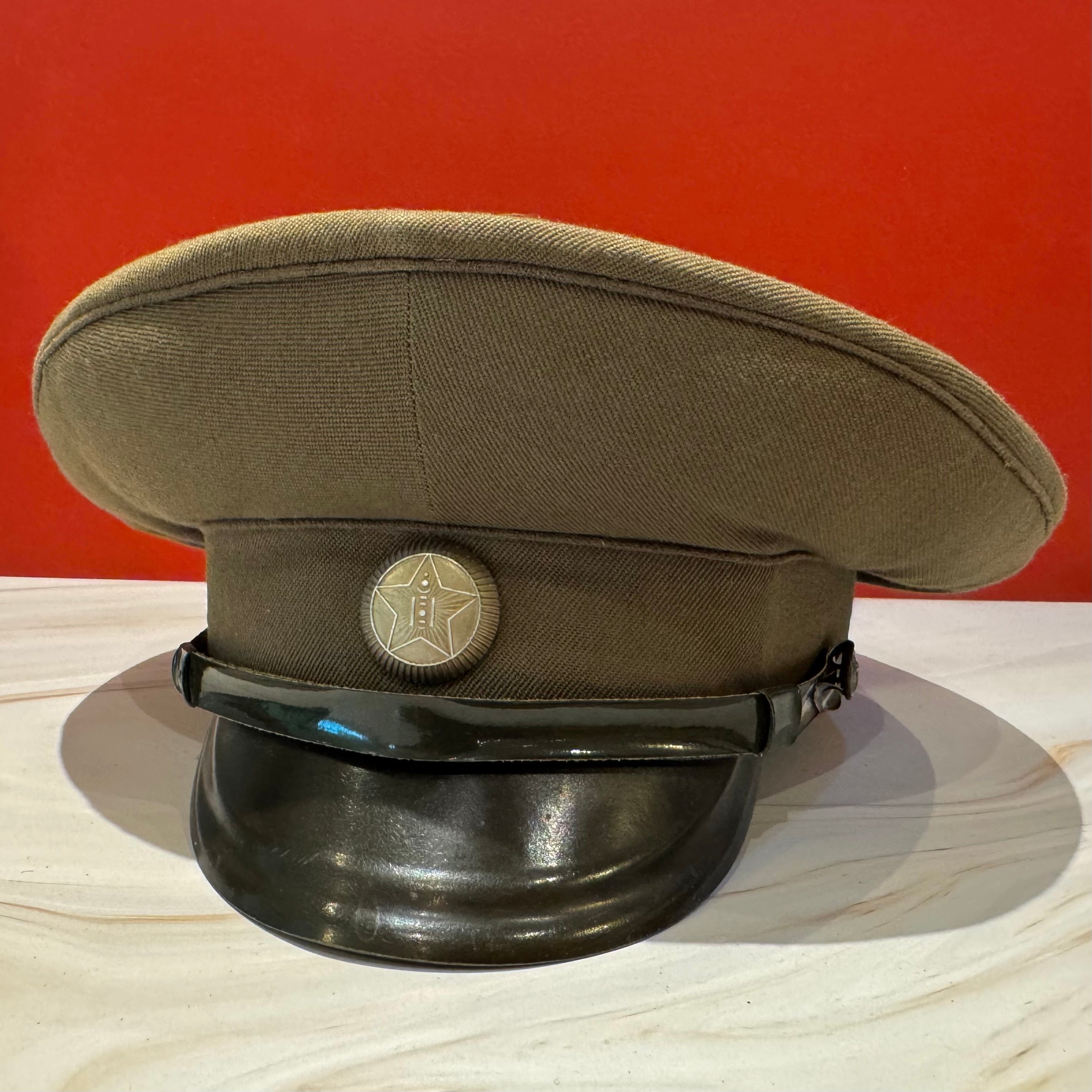 1970s Mongolian People's Army field officer visor cap with olive wool crown, black chin strap, and metal MPA star insignia. Represents Cold War-era officer gear, reflecting the transitional styling between parade and combat dress.