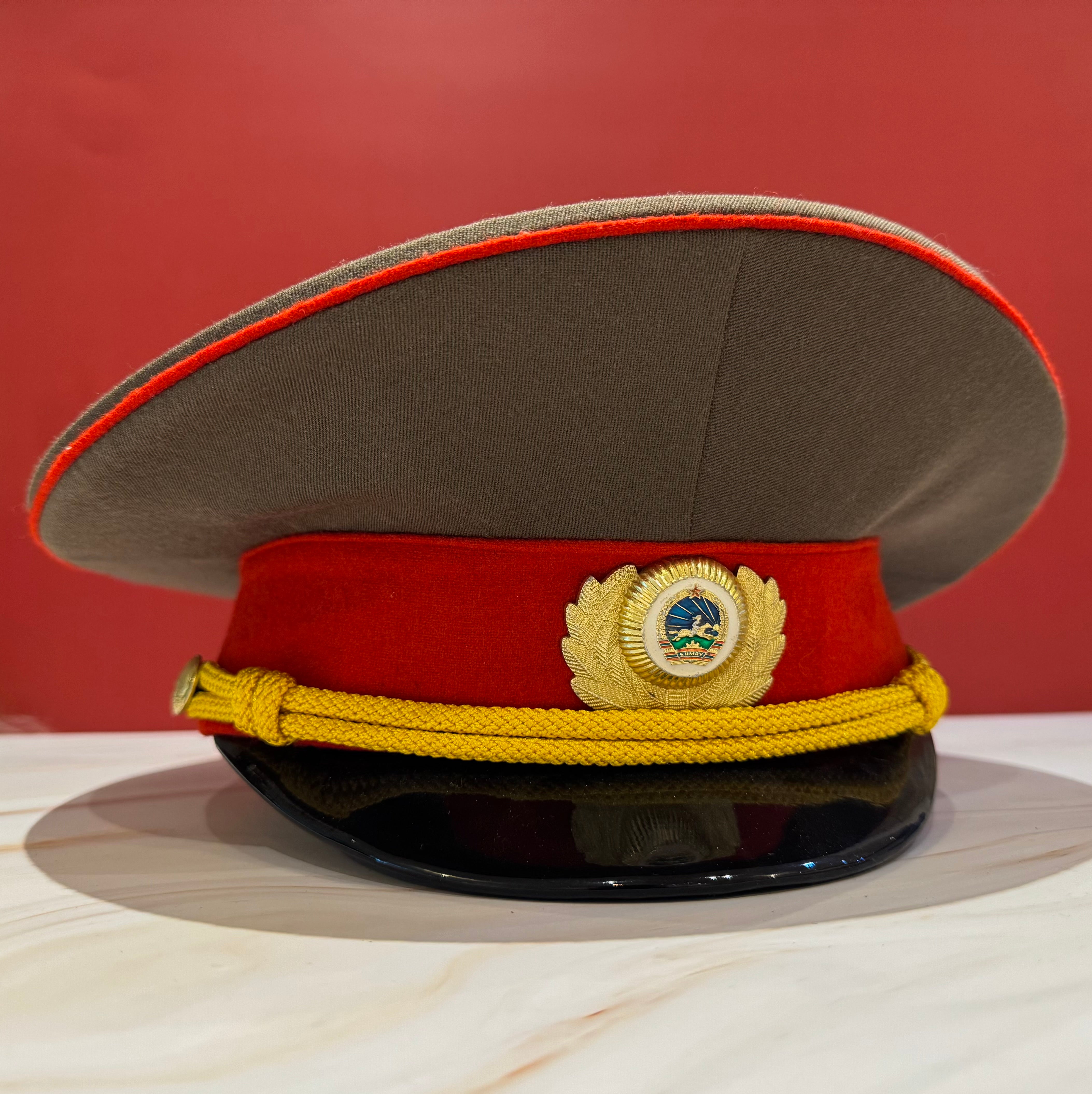 1980s Mongolian People's Army general rank service visor cap in near mint condition, featuring red band with gold bullion chin cord, national cockade, and black polished visor. Represents Cold War-era military aesthetics with socialist-era design standards.